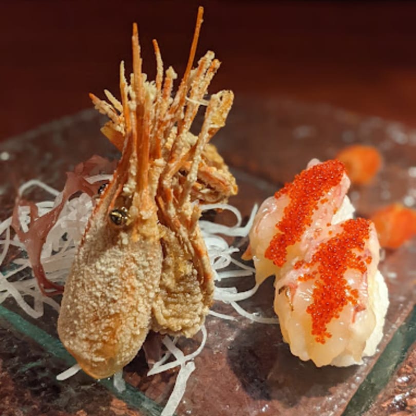 Crispy fried shrimp heads and two pieces of sushi topped with red roe on a glass plate.