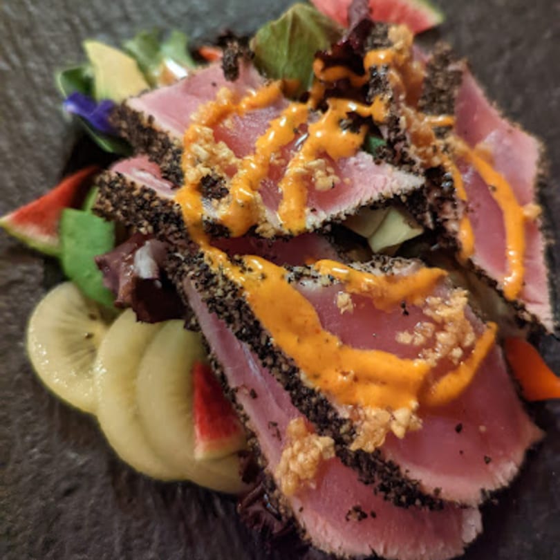 Seared tuna slices with black pepper crust, orange sauce, and fruit on a dark plate.