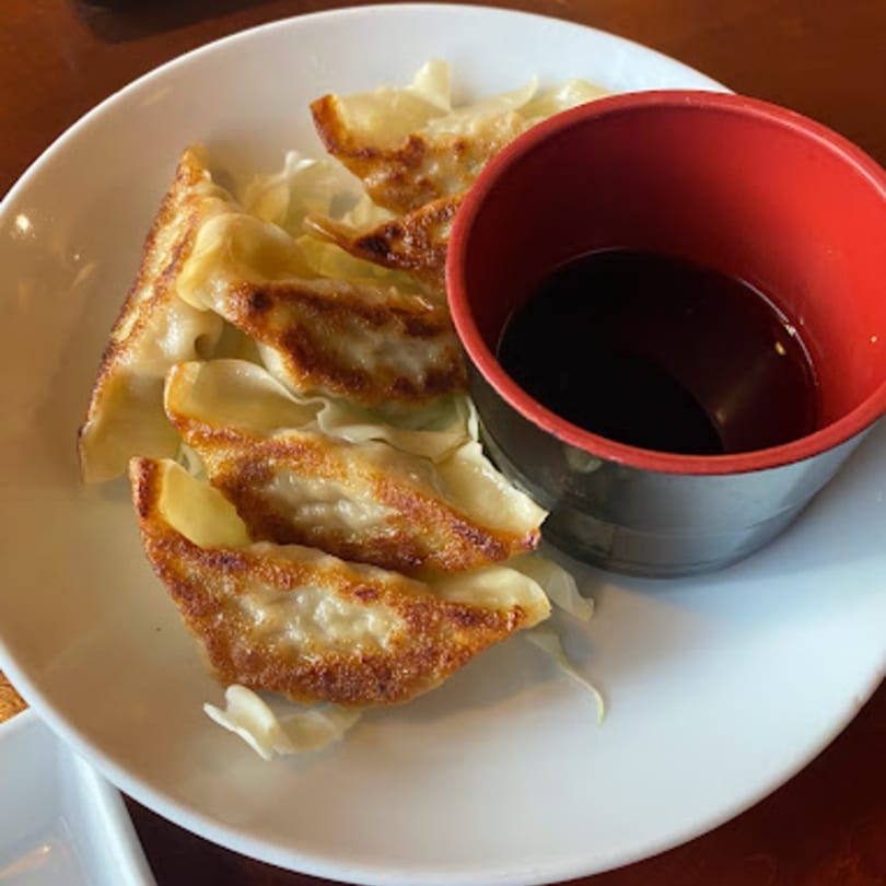 Golden-brown Mikado Ryotei gyoza dumplings with a side of soy sauce in a red and black bowl.