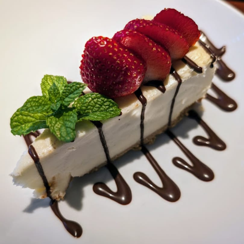 A slice of cheesecake with strawberries, mint, and chocolate drizzle on a white plate.